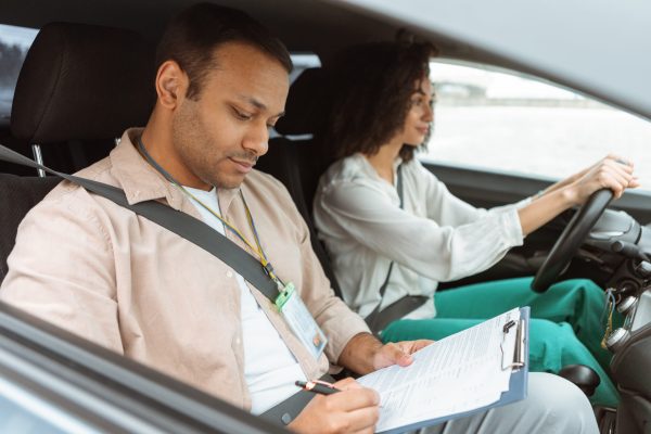 Driving Test. Arabic Instructor Man Taking Notes In Lesson Plan Teaching Novice Driver Woman Sitting In Car. Coach Mastering And Evaluating Learner's Practical Skills. Selective Focus, Side View