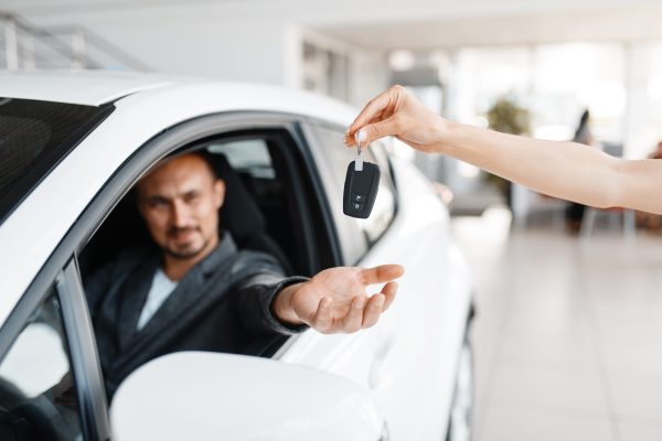 Happy man takes the key to the new car in showroom. Male customer buying vehicle in dealership, automobile sale