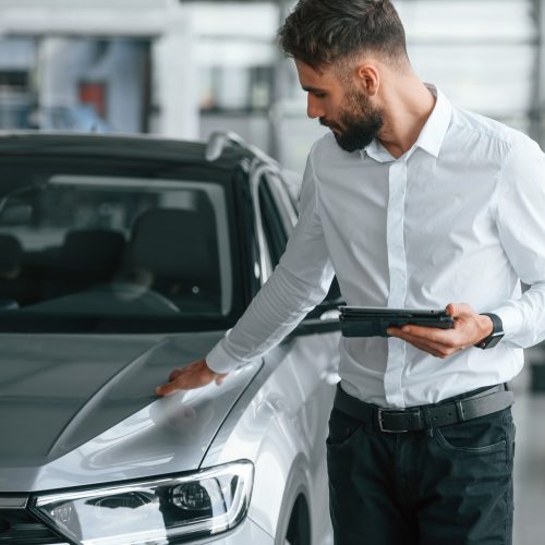 Touching the surface of automobile. Young man in white clothes is in the car dealership.